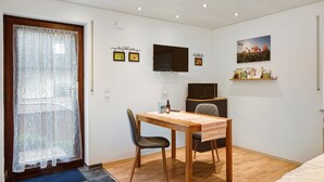 A small, well-lit room with a wooden dining table, a single chair, a bed, a TV mounted on the wall, and a door with a sheer curtain. - Studio 'Gelis' with Mountain View, Private Terrace and Wi-Fi (Ihringen)