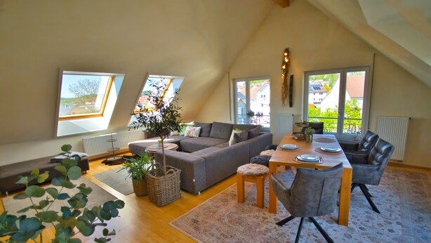 Un espacio tipo loft con sofá, mesa de comedor, sillas, ventanas amplias y una planta.