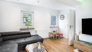Ein modernes Wohnzimmer mit einem grauen Sofa, Holzboden, einem Fernseher und einem Fenster mit Blick auf Gebäude.