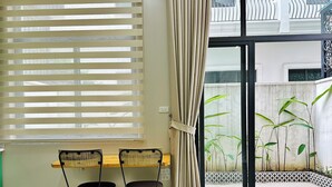 A room with a window covered by blinds, a curtain, a small table with two chairs, and a view of a garden through the window. - Anvi House - Vinhomes Marina (Hai Phong)