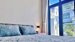 A bedroom with a large bed, a nightstand, and a view of a building through a glass door. - Anvi House - Vinhomes Marina (Hai Phong)