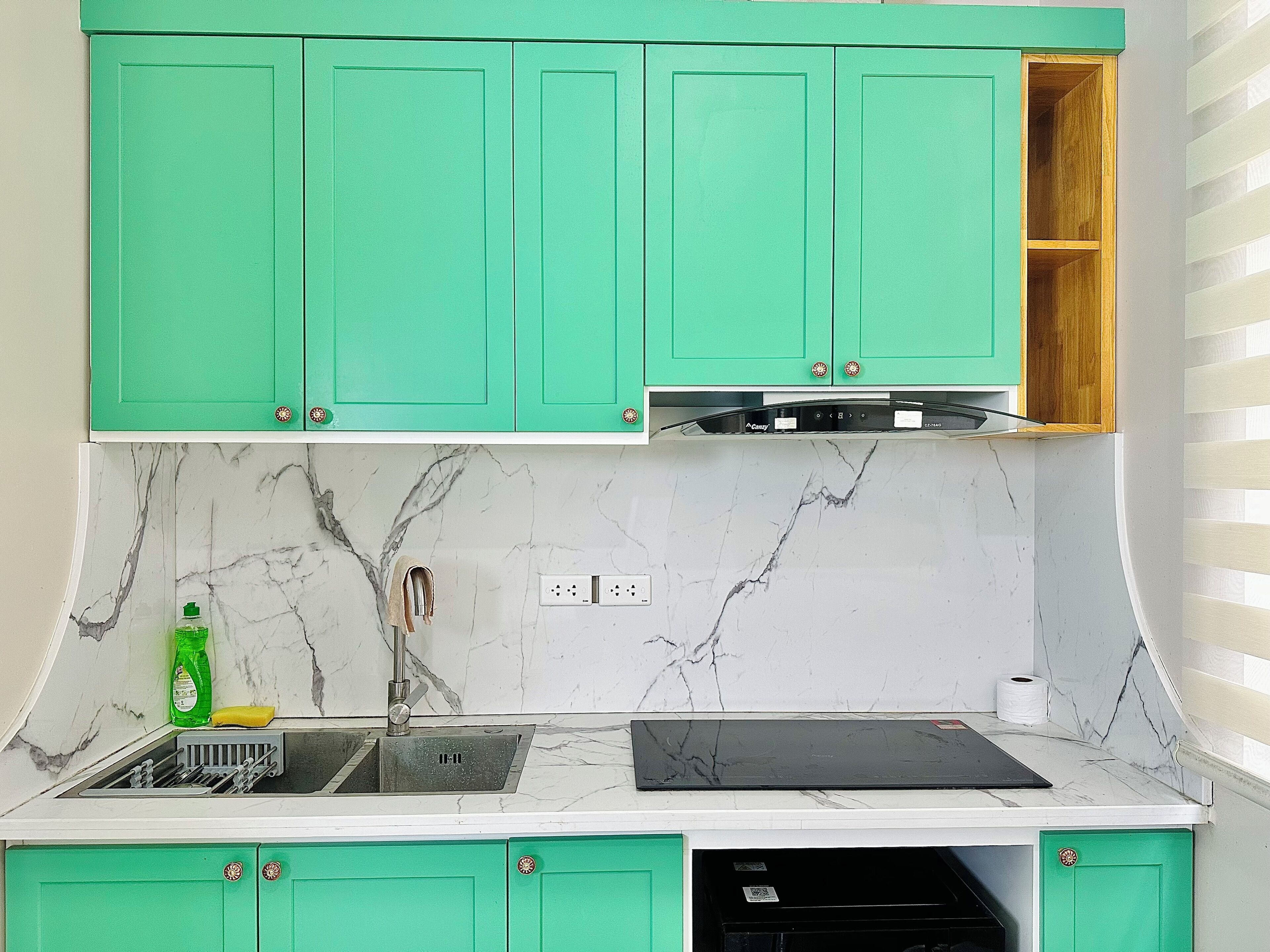 A kitchen with green cabinets, a marble backsplash, a sink with dish rack, and a black countertop.