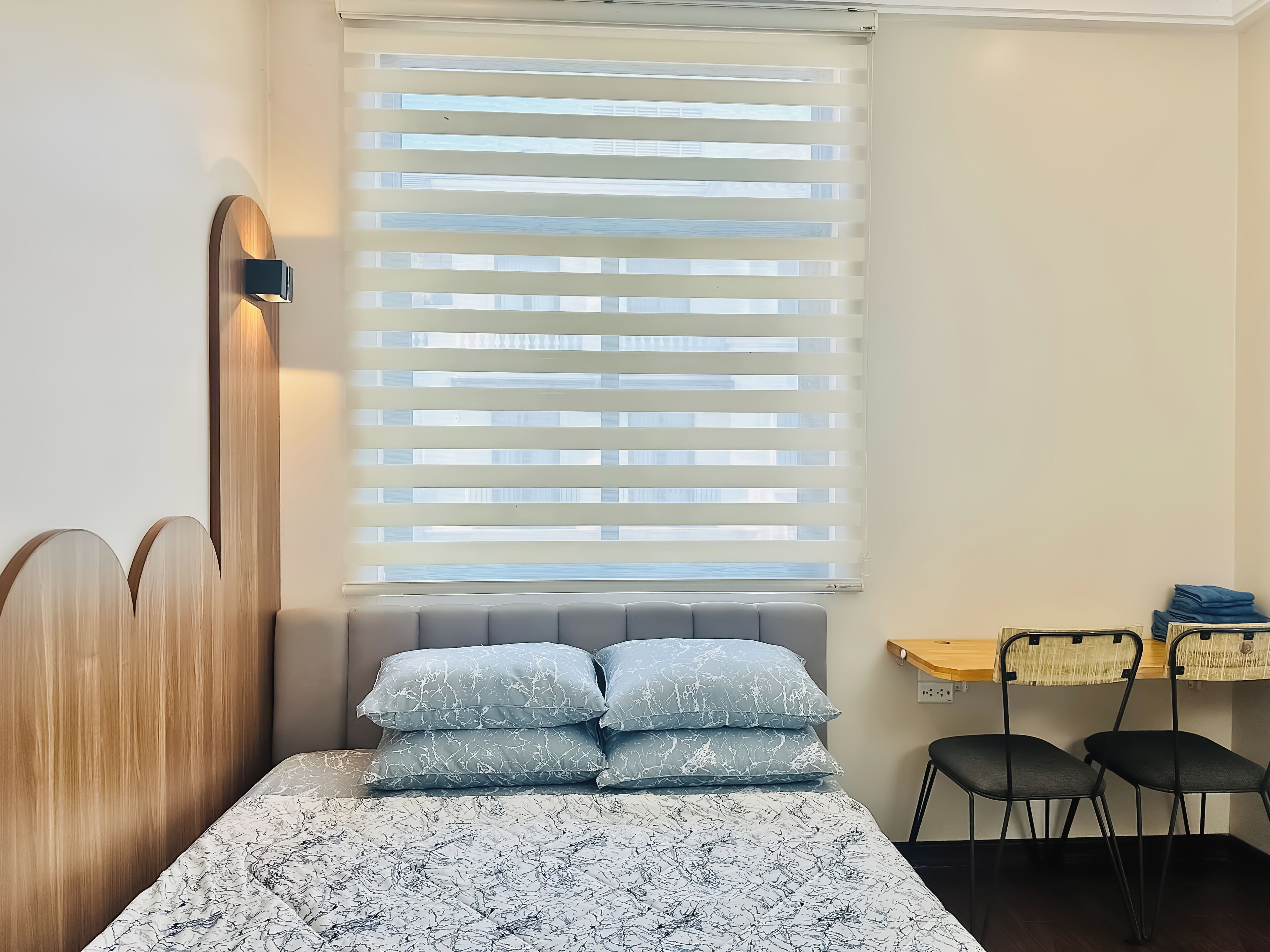 Un dormitorio con cama, cabecera, ventana con persianas, mesita de noche y dos sillas.