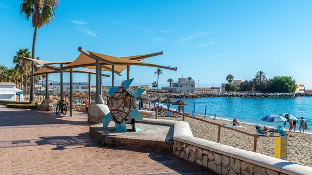 Un paseo marítimo con una palmera, un área de descanso cubierta y vistas al mar.