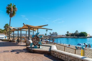 A beachside promenade with a palm tree, a covered seating area, and a view of the sea.