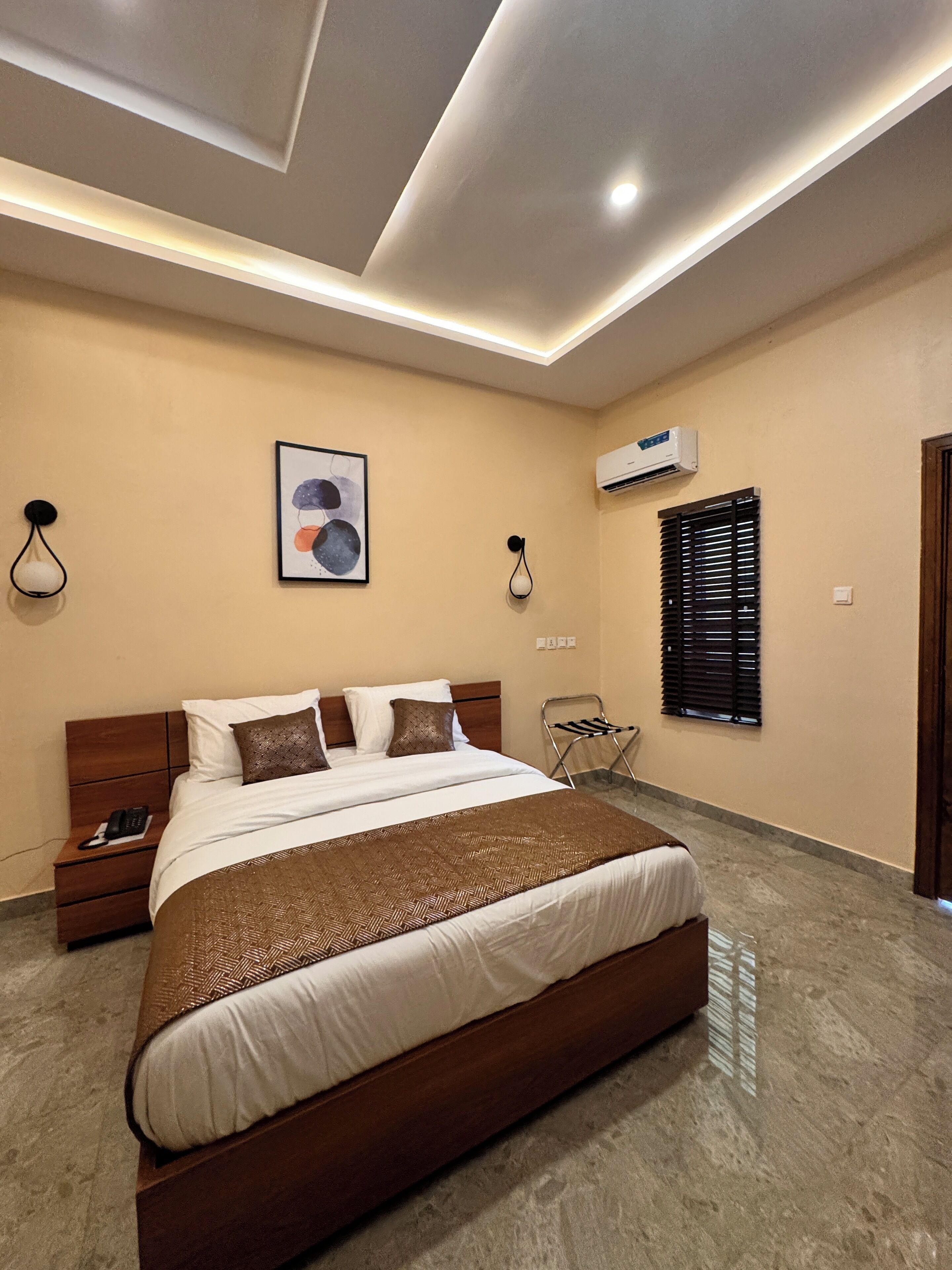 A modern bedroom with a large bed, a nightstand, a wall-mounted air conditioner, and a window with blinds.