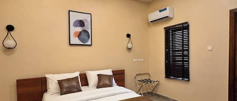 A modern bedroom with a large bed, a nightstand, a wall-mounted air conditioner, and a window with blinds.