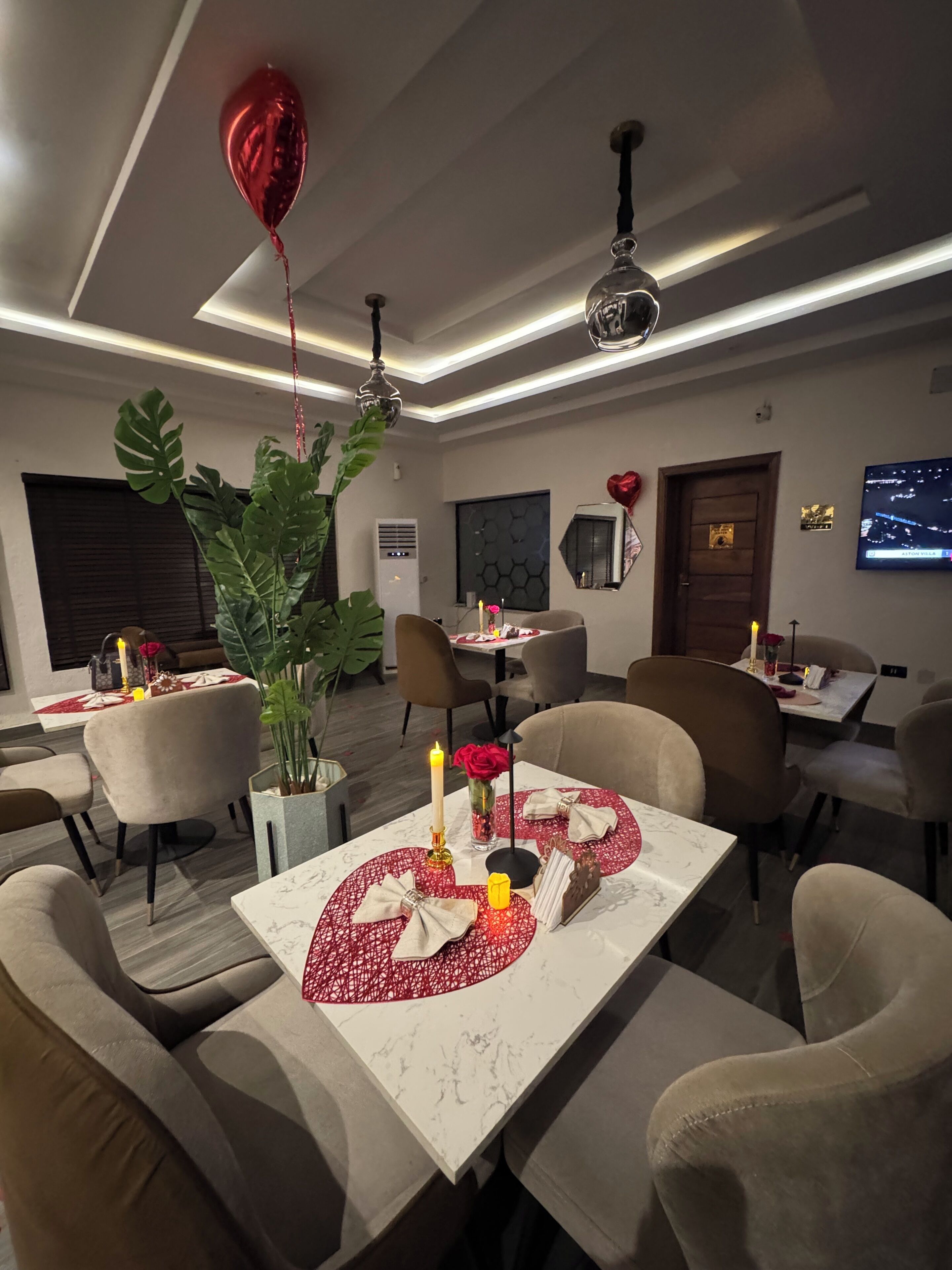 A dining area with tables set for two, featuring red table runners, candles, and a potted plant.
