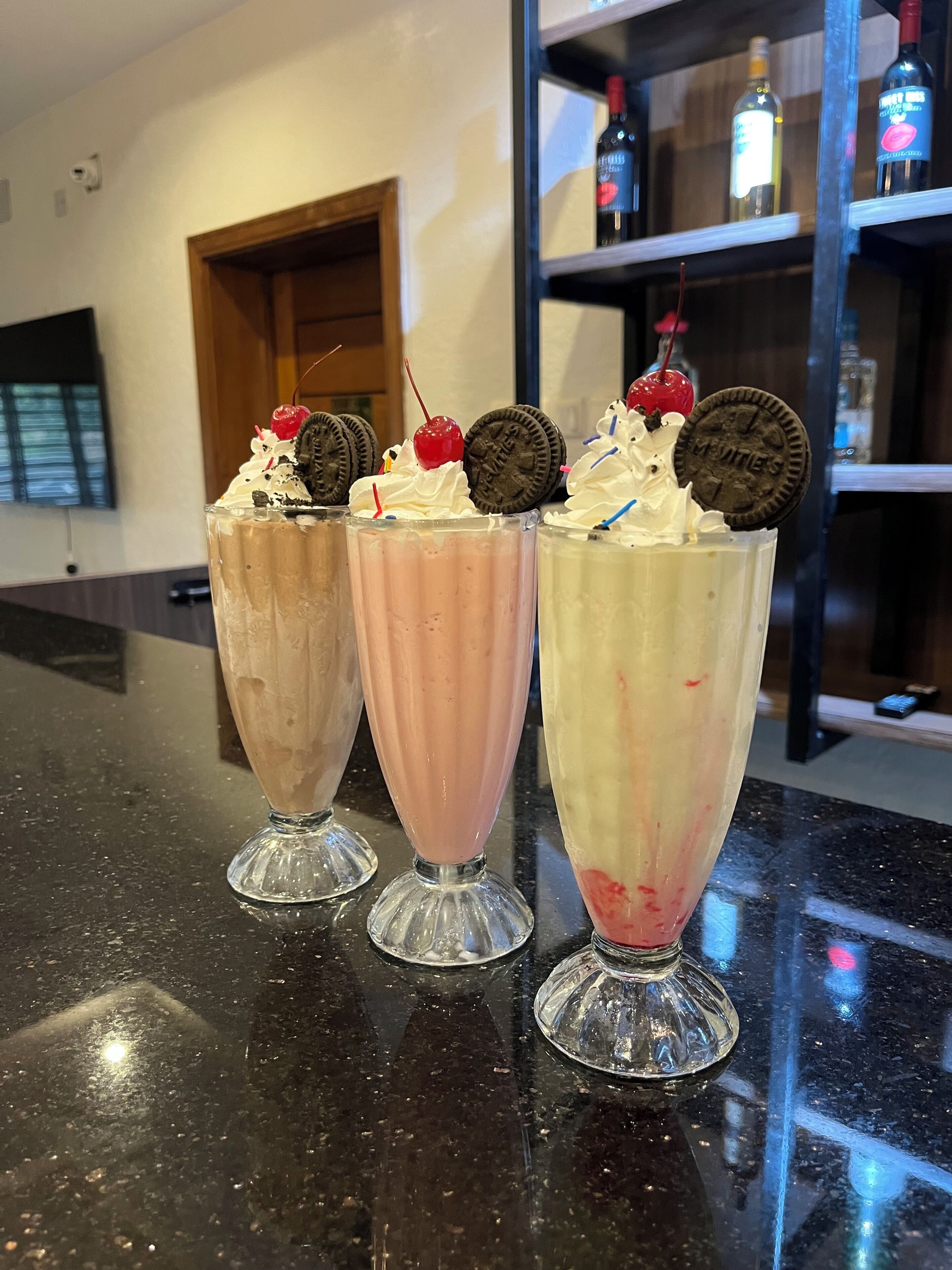 Three milkshakes on a counter with a wooden door and shelves in the background.