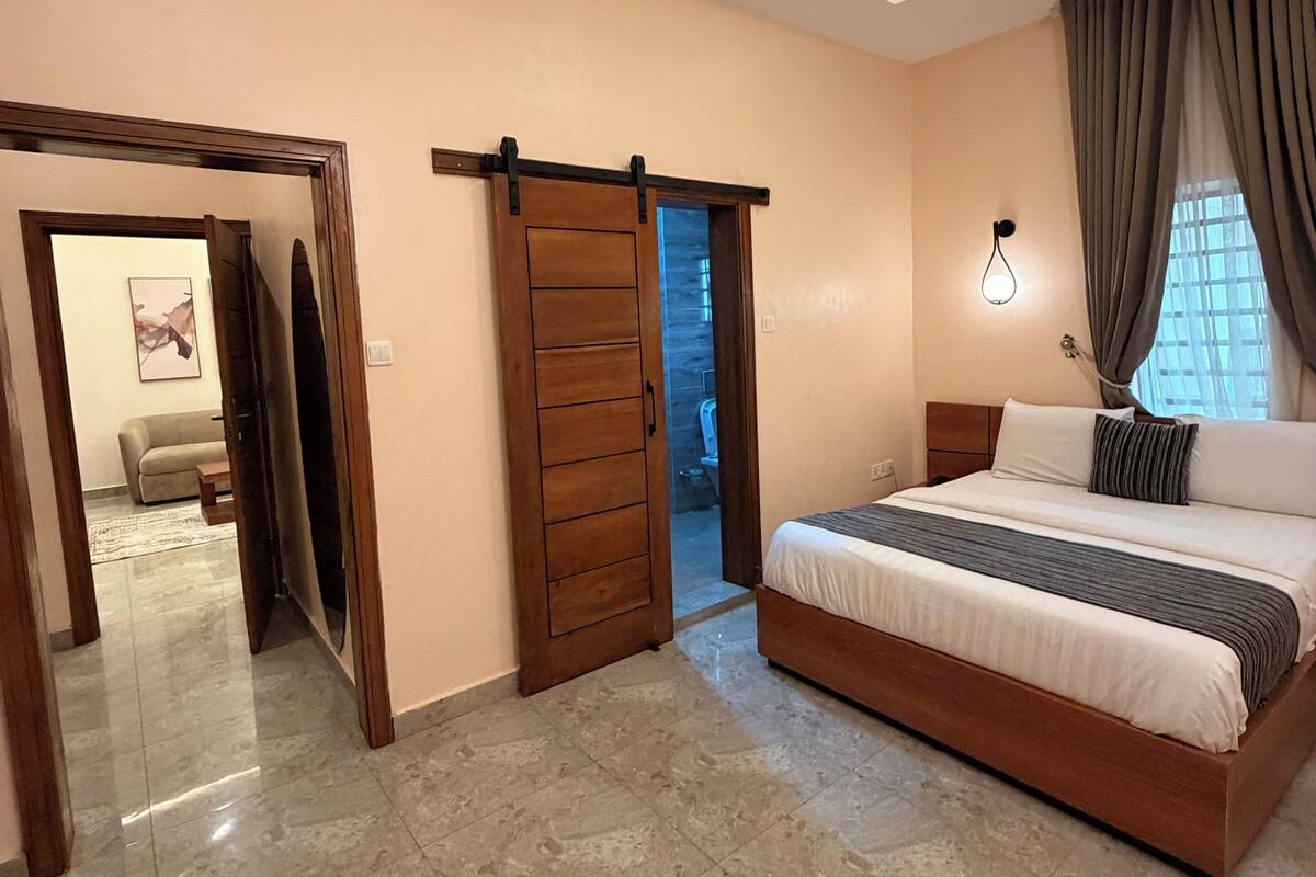 A bedroom with a bed, a sliding door, a wall-mounted light, and a view through a window.