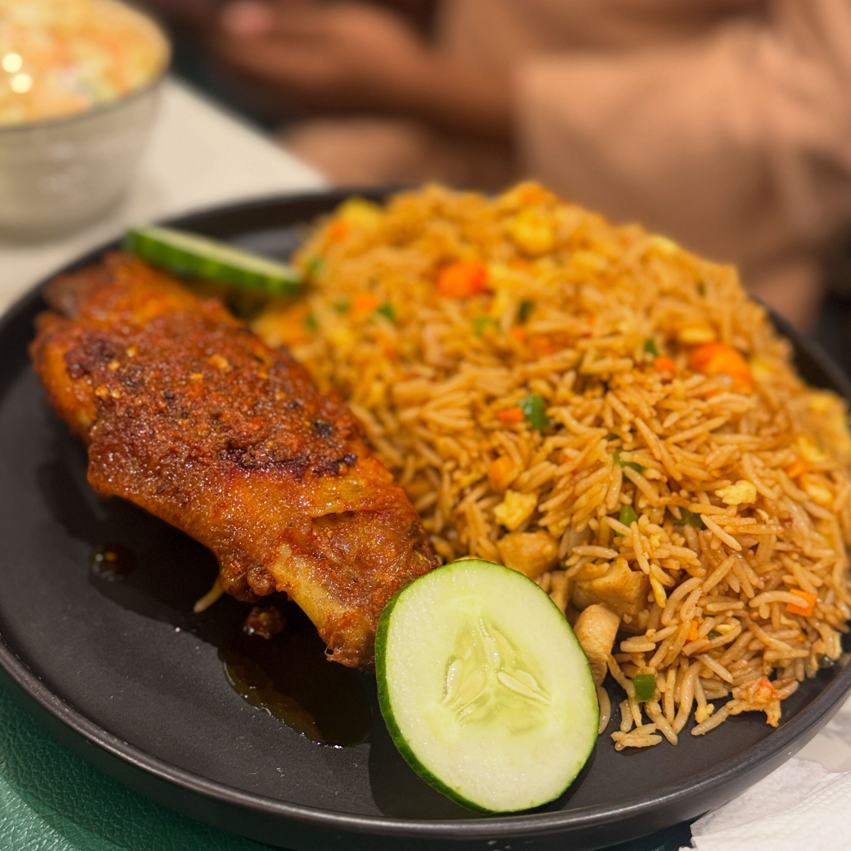 A plate of fried rice with vegetables and a piece of fried chicken, served on a black plate with a slice of cucumber.