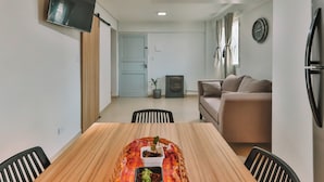 A modern living room with a wooden dining table, a sofa, a TV mounted on the wall, and a small potted plant.