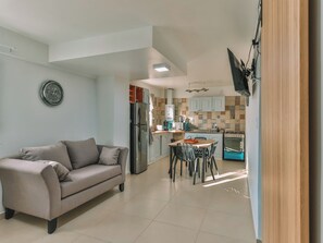 A modern living room with a sofa, a dining area with chairs, a kitchen with cabinets and appliances, and a TV mounted on the wall.