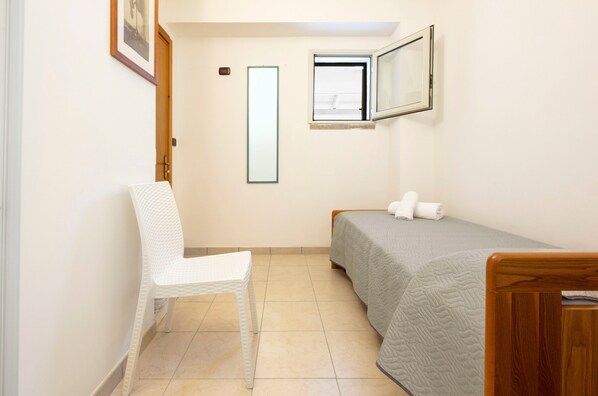 Un dormitorio pequeño y ordenado con una cama individual, una silla, una mesita y una ventana.