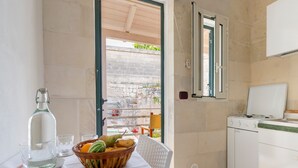 Una cocina con estufa, refrigerador, ventana con vista a un muro de piedra, y una mesa con frutero y vasos.