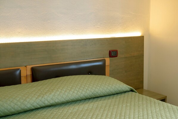 Una cama con cubrecama verde acolchado, cabecera de madera y mesita de noche con un reloj rojo.