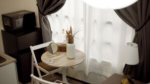 A compact hotel room with a small dining table, a white chair, a microwave, a small cabinet, a bedside table with a lamp, and a window with curtains. - RUTiLE IKEBUKURO TOKYO (Tokyo)
