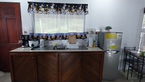 A compact kitchen with a wooden cabinet, a coffee maker, a refrigerator, a sink, and a window with a patterned curtain.