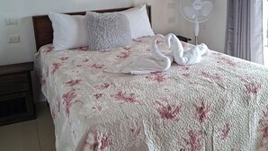 A neatly made bed with a floral bedspread and two pillows, a standing fan, and a wooden ceiling.