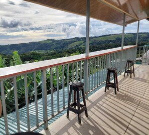 A covered outdoor area with a view of rolling hills, a railing, and two stools. - Alan's House (Santa Elena)