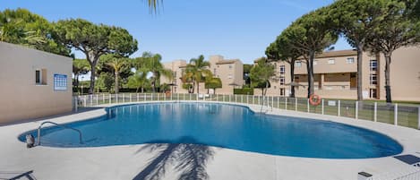 A swimming pool with a white fence, palm trees, and apartment buildings in the background.