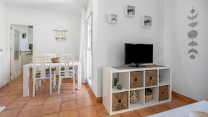 Ein modernes Wohnzimmer mit einem weißen Fernsehschrank, einem Esstisch mit Stühlen und einer Küchenzeile im Hintergrund.