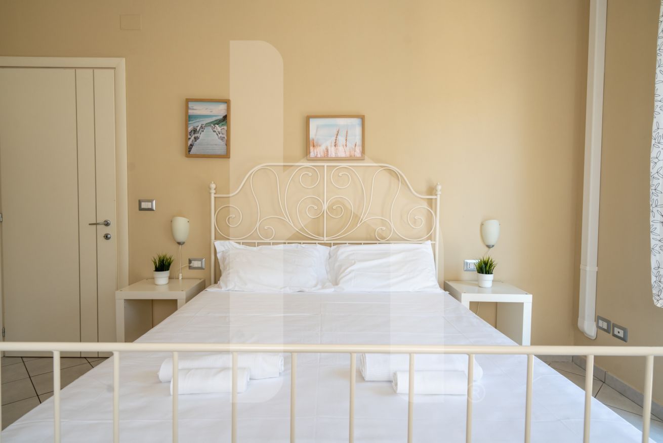 Une chambre avec un lit en métal blanc, deux tables de chevet avec des lampes et une porte sur le côté gauche.