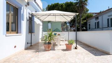 Un'area salotto esterna coperta, con tavolo e sedie, piante in vaso e un ombrellone bianco.