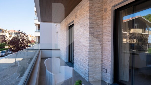 A modern balcony with white wicker furniture, a stone wall, and a glass door leading to a covered area. - Homiday - Residence Pinetina Int. 02 (Pineto)