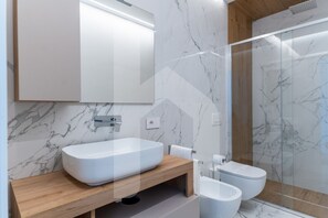 A modern bathroom with a marble backsplash, a wooden vanity, a freestanding sink, a toilet, and a glass shower enclosure. - Homiday - Pinetina Residence Int. 05 (Pineto)