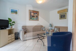A living room with a beige sofa, a glass coffee table, a blue armchair, a television on a cabinet, and framed pictures on the walls. - In the heart of the Torre del Cerrano Marine Protected Area, a few steps from the beach is the Green Bay Village. The three-room apartment A01, on the mezzanine floor of the GRAN SASSO building, has an independent entrance with a small garden, pool view, (Pineto)