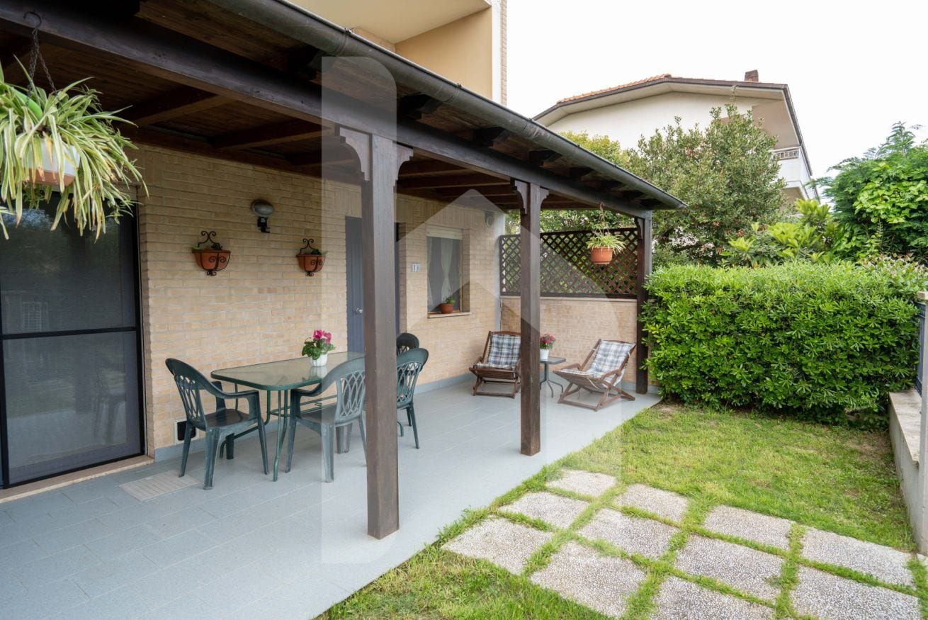 Un patio techado con una mesa y sillas, plantas en macetas y una vista de una casa al fondo.