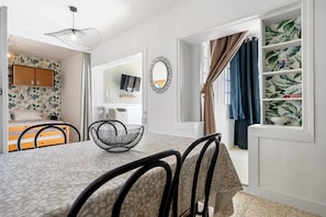 Una stanza con un letto, una TV, un tavolo da pranzo con sedie, una mensola con oggetti decorativi e una finestra con tende.