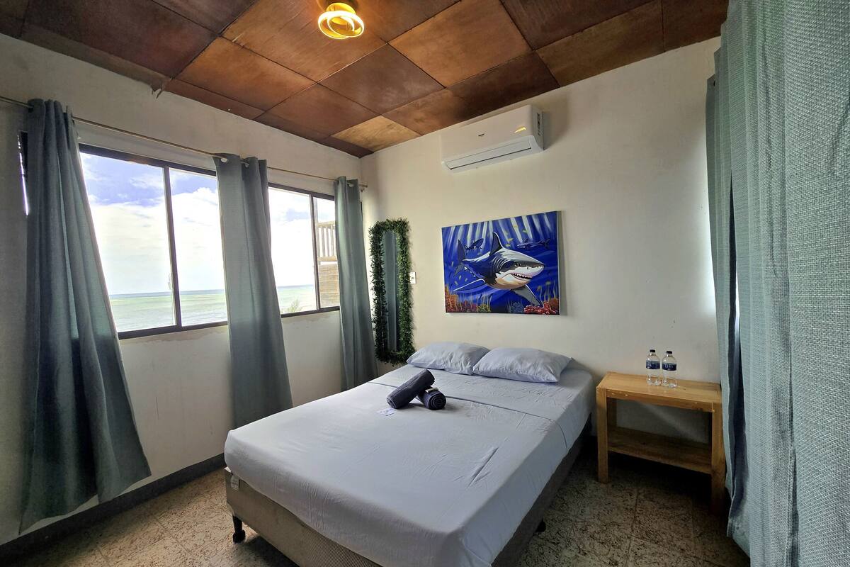 A bedroom with a bed, a window with curtains, an air conditioning unit, a painting of a shark, and a nightstand with bottles.