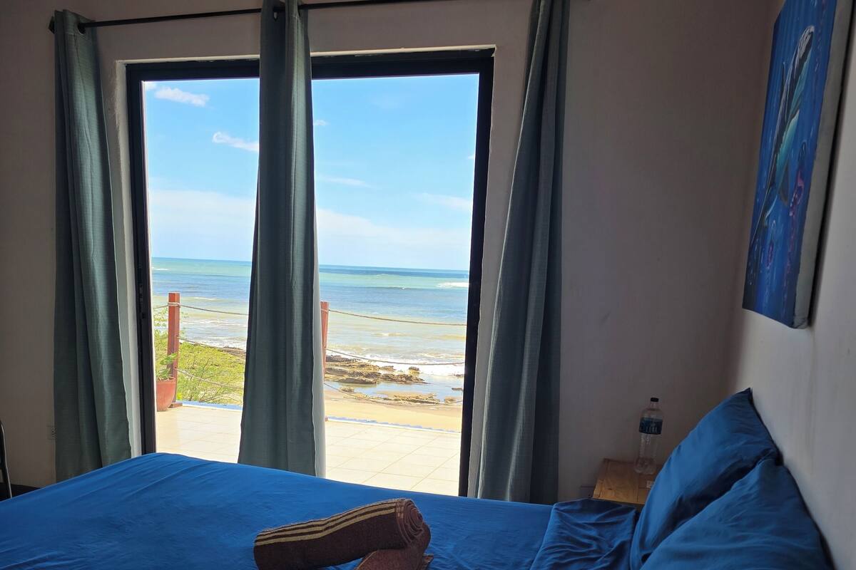 A bed with blue bedding, a rolled towel, and a pillow, with a view of the beach and ocean through a window.