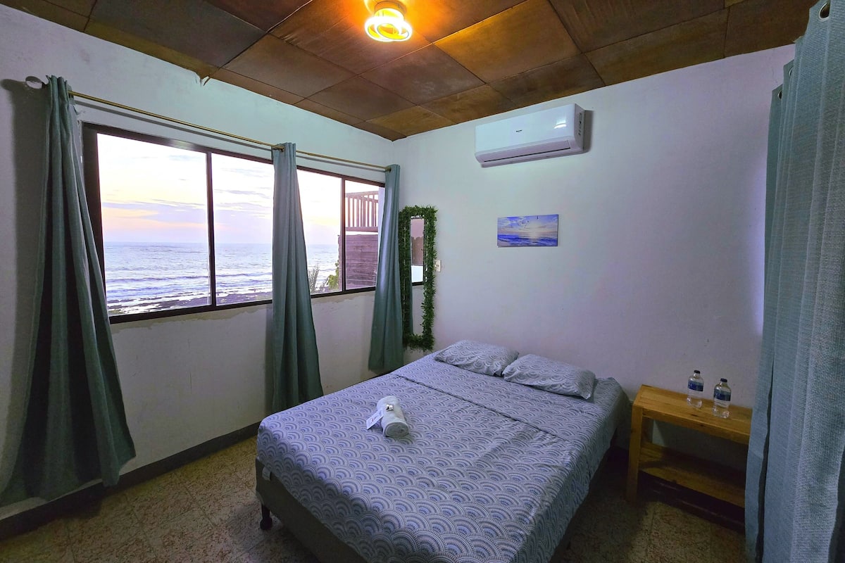 A bedroom with a bed, a window with curtains, a wall-mounted air conditioner, a small bedside table with water bottles, and a painting on the wall.