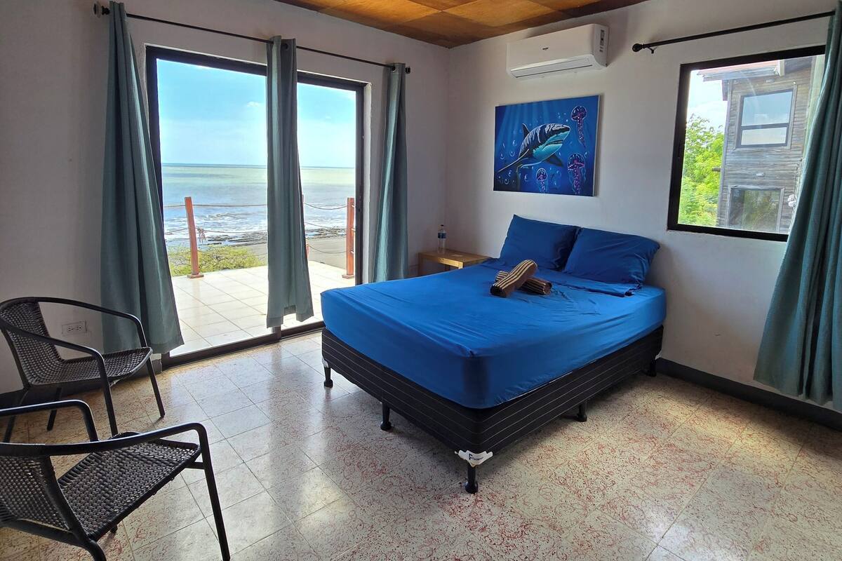 A bedroom with a bed, a chair, a painting, and a view of the sea.