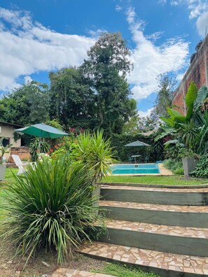 A pool area with steps leading down to the water, surrounded by lush greenery and a clear blue sky.