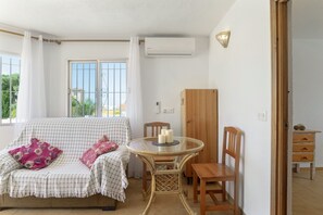 Une pièce avec un canapé à carreaux, une table ronde en verre ornée de bougies, une chaise en bois, un meuble en bois et une fenêtre donnant sur un paysage verdoyant.