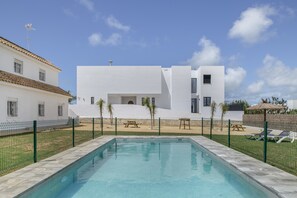 Une piscine entourée d’une clôture, un bâtiment blanc doté d’un toit plat et un ciel d’un bleu limpide.