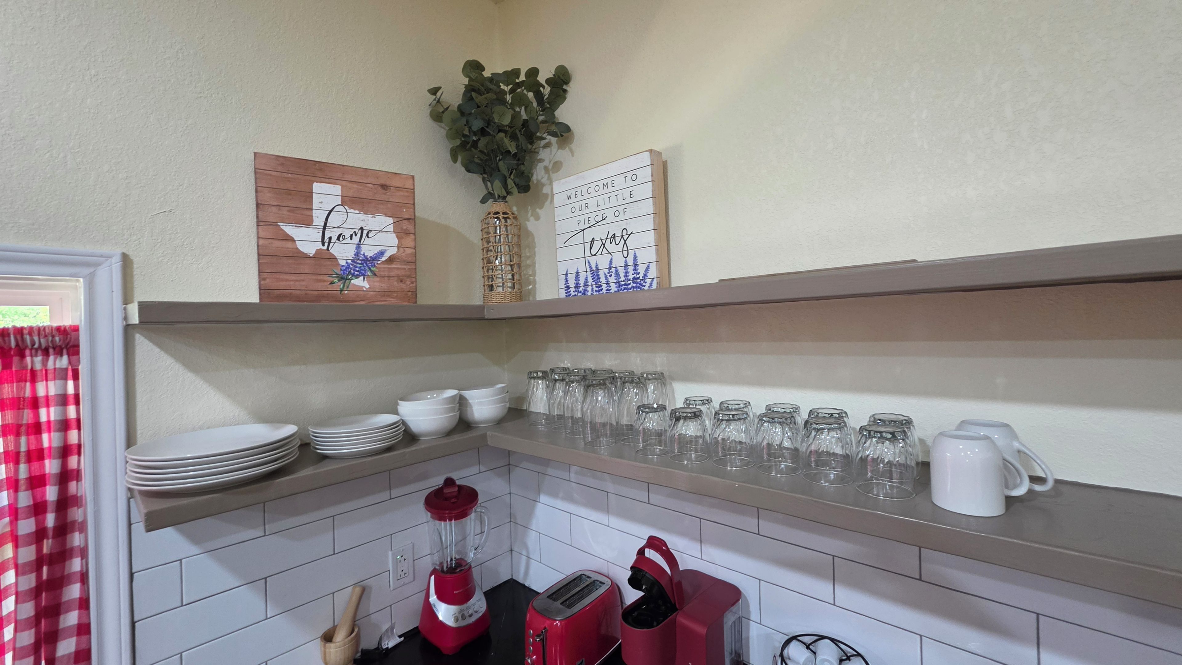 A kitchen with white tiled walls, a red toaster, a blender, and a coffee maker. There are shelves with plates, bowls, and glassware.