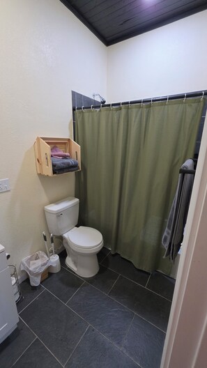 A bathroom with a toilet, a shower curtain, a wooden shelf with towels, and a floor with dark tiles. - 3-bedroom house in charming Waco with WiFi, AC. Start your getaway (Waco)