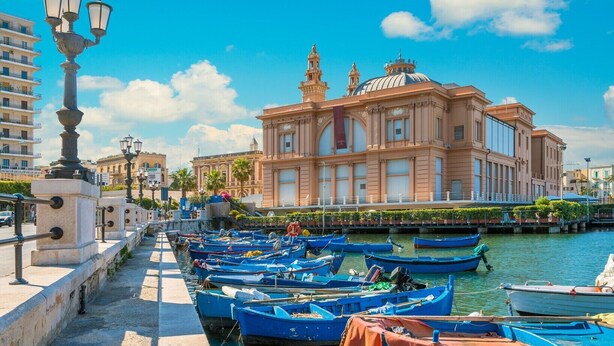 A waterfront with boats docked, a prominent building with classical architecture, and a clear blue sky.