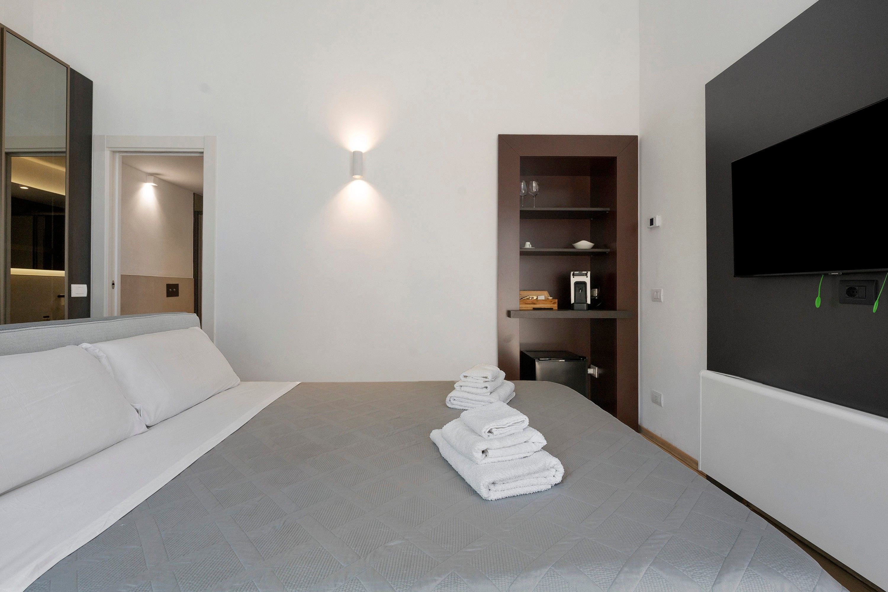 Habitación de hotel moderna con una cama grande, un televisor de pantalla plana montado en la pared, un mueble de almacenamiento empotrado y un espejo en la pared izquierda.