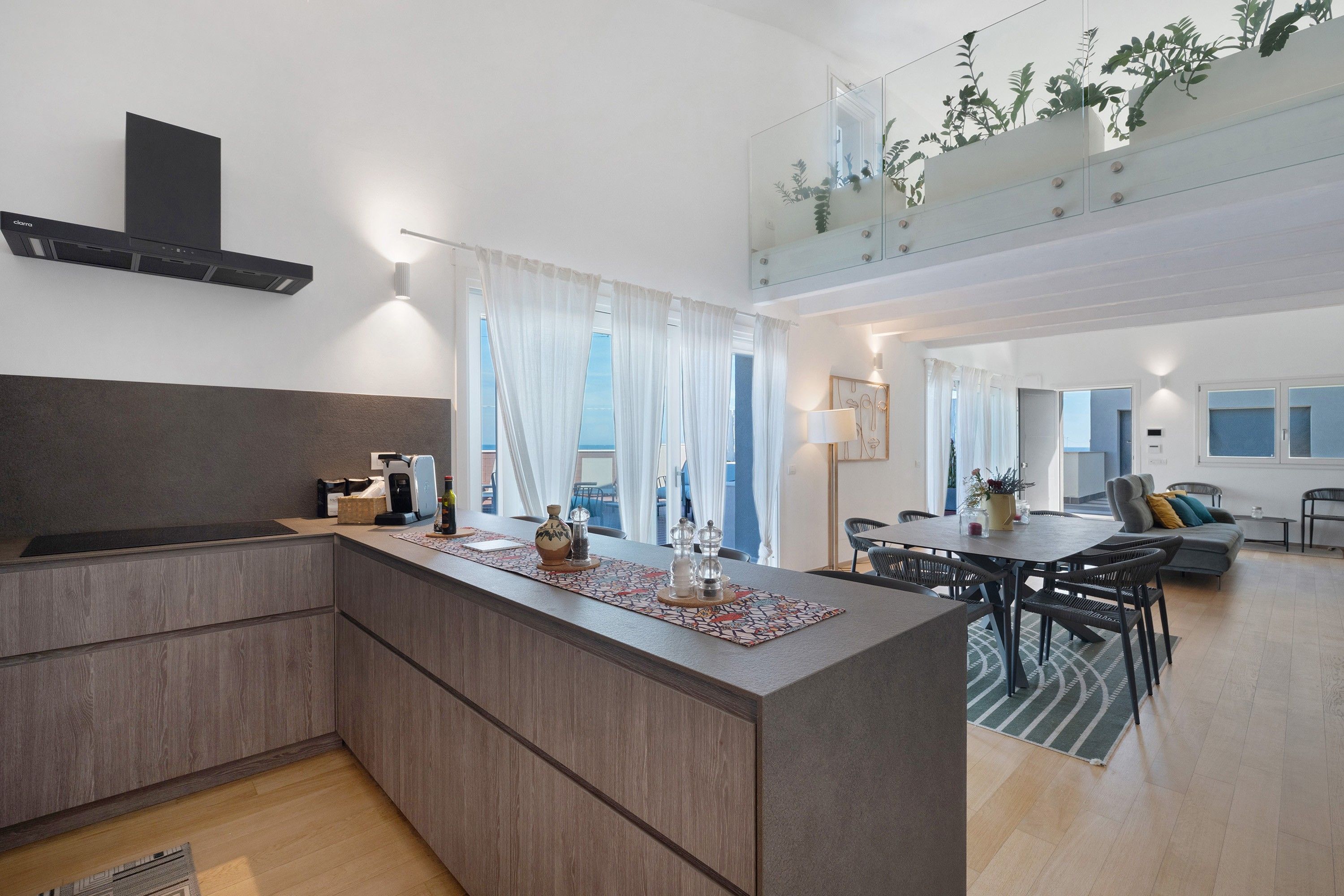 Una cucina moderna con un'isola centrale, un'area pranzo con tavolo e sedie, e un soggiorno con divano e un balcone con piante.