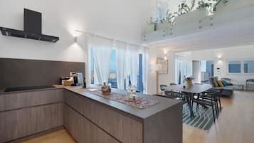 Una cucina moderna con un'isola centrale, un'area pranzo con tavolo e sedie, e un soggiorno con divano e un balcone con piante.