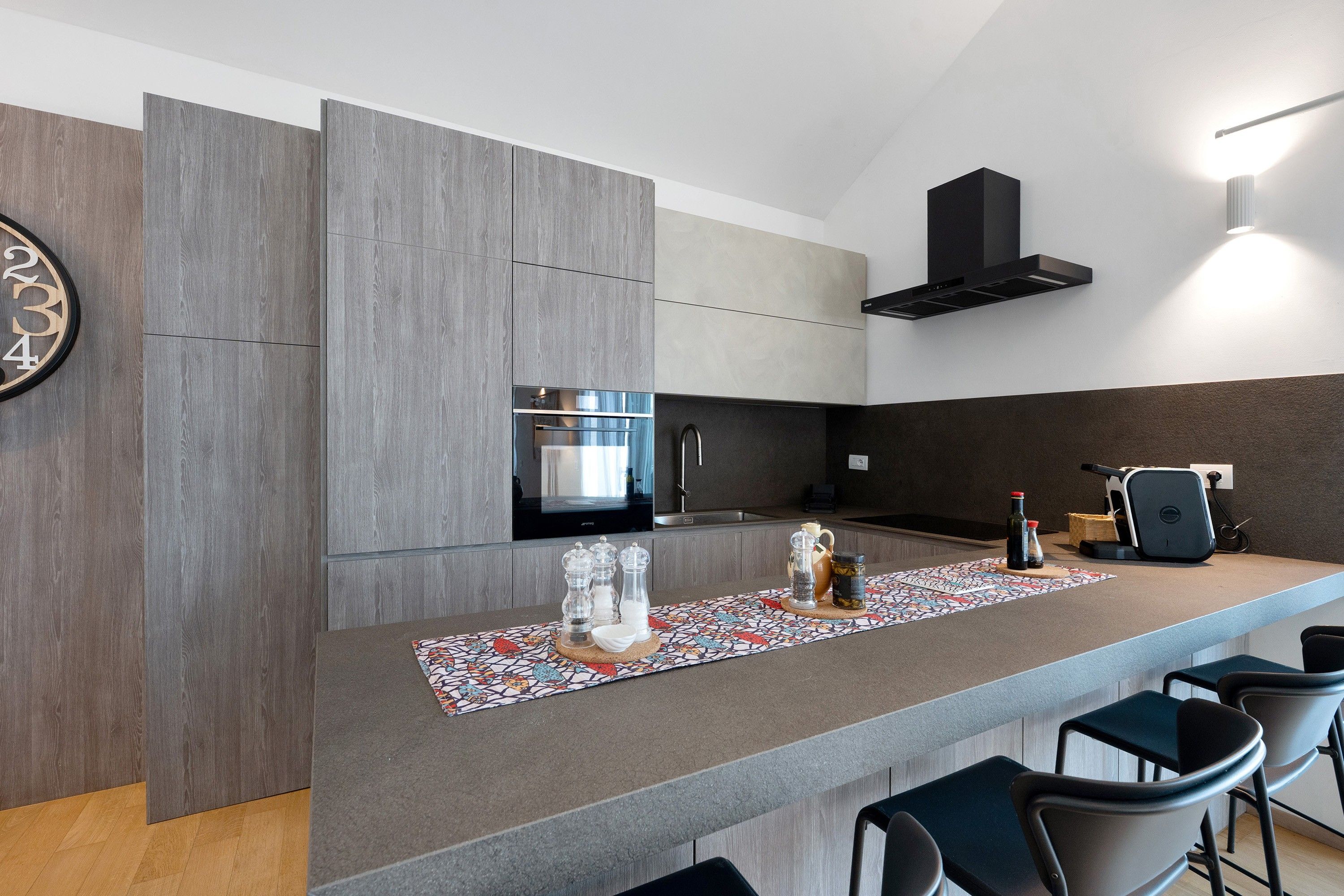 Une cuisine moderne équipée d’un four encastré, d’un four à micro-ondes et d’une cuisinière à gaz. On y trouve également un coin bar avec un chemin de table à motifs et des tabourets de bar.