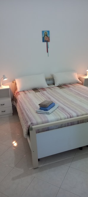 A bedroom with a bed, two bedside tables, a nightstand, a wall-mounted picture, and a tiled floor.