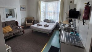 A compact hotel room with a bed, a sofa, a small table, a kitchenette, and a window with curtains.