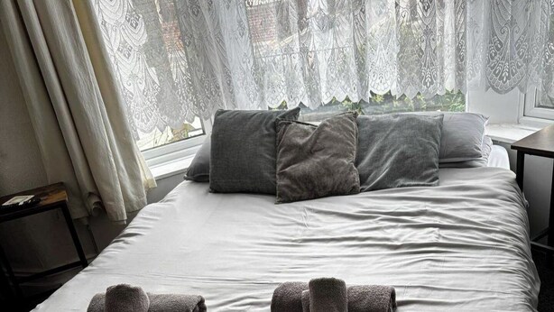 A neatly made bed with grey pillows and towels, lace curtains, and a view of greenery outside.